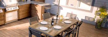 A large wooden deck with a grill on one side and lounge chairs on the opposite side. In the middle is a six-person dining table with a large green umbrella hanging over it.