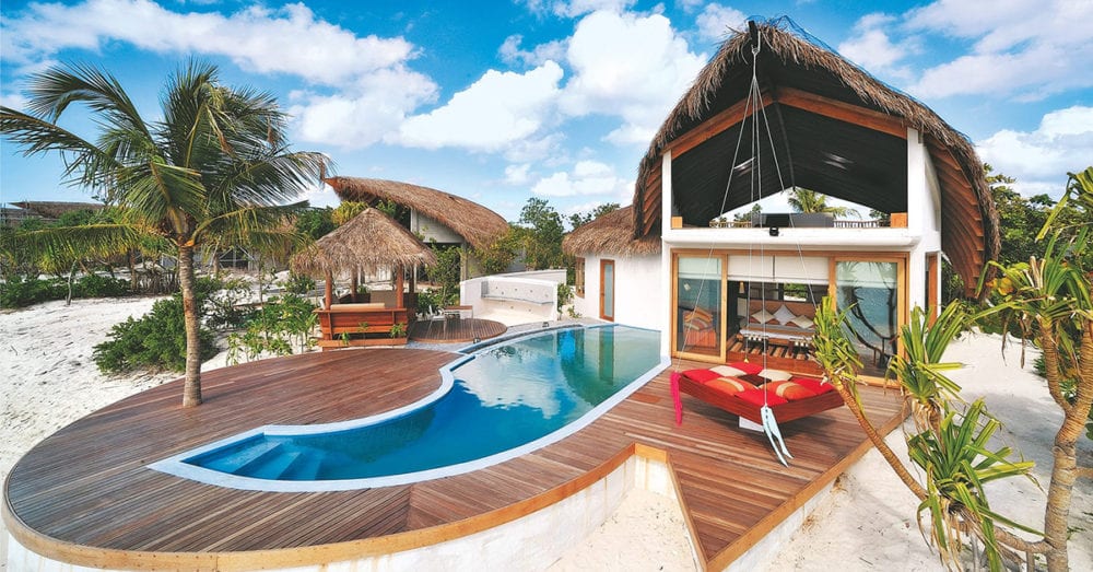 This image depicts a beachfront wooden deck with a pool, a hanging bed and a cabana next to it, surrounded by palm trees.