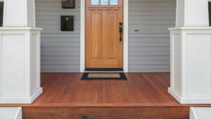 A front porch of a home with gray siding. The deck features red douglas fir wood, a wooden front door, and large white columns.