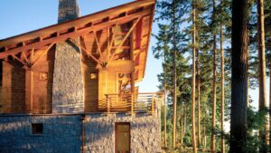 A home with an asymmetrical roof, cedar siding, as well as a gray stone chimney and first level. The porch overlooks a forest of pine trees.