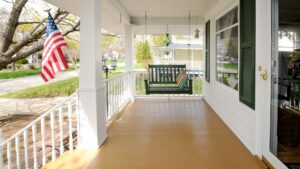 A covered porch of a white home with green shutters and an American flag. A swinging bench hangs above the Douglas Fir porch flooring.