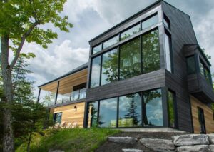This image depicts a two-story house with both black and natural wood siding. Large windows reflect a tree. 