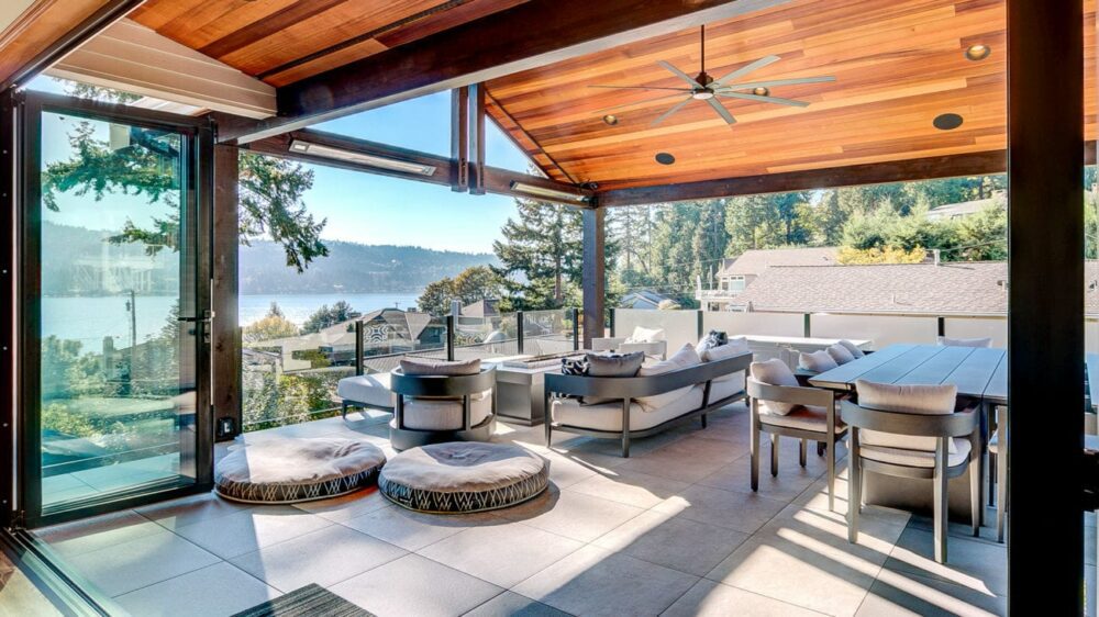 This image depicts a modern indoor-outdoor patio with gray tiles, glass railings and a wooden gable roof with a large black fan. The patio is furnished with a matching, black, white and gray-themed dining table and fireplace area surrounded by couches. The patio overlooks a blue lake.